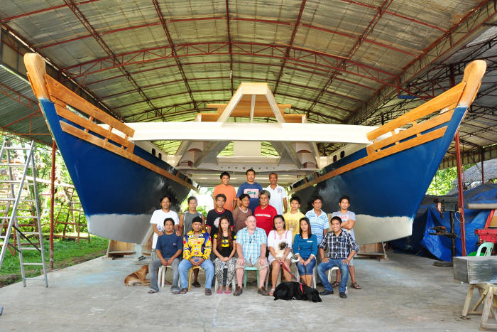 A team of boat builders with an almost-finished large blue catamaran under a shelter