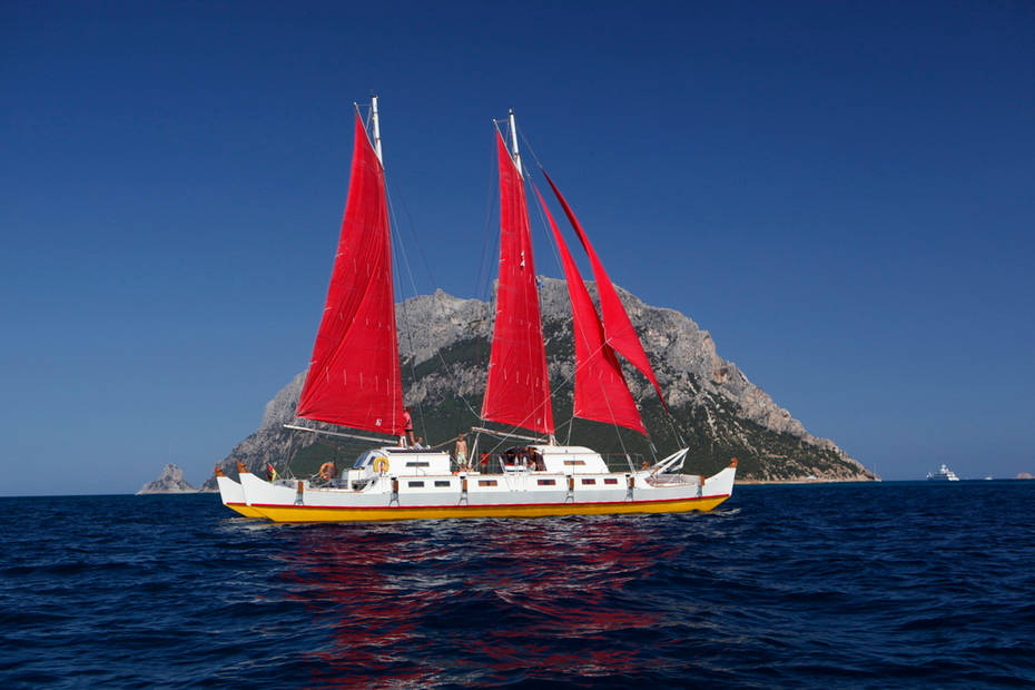 White and red catamaran, sailing against a backdrop of a rocky island
