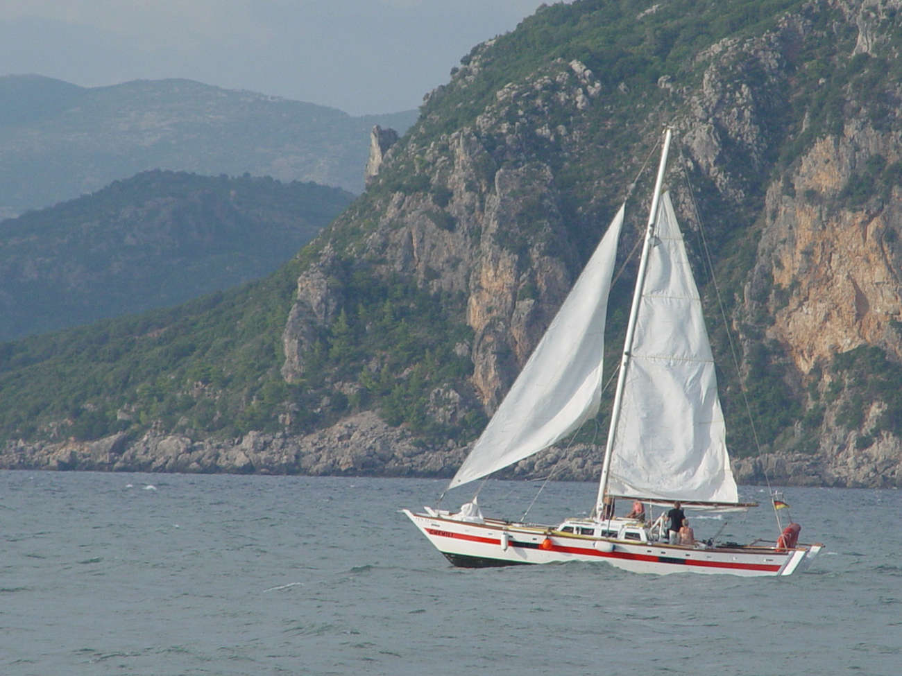 Red and white Narai, two people aboard, sailing against a backdrop of mountains covered in greenery