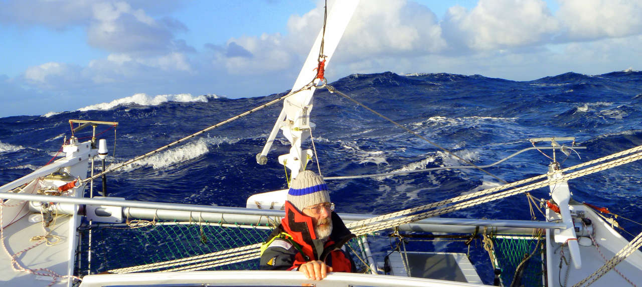 A huge wave approaching the stern of a catamaran, one man at the tiller
