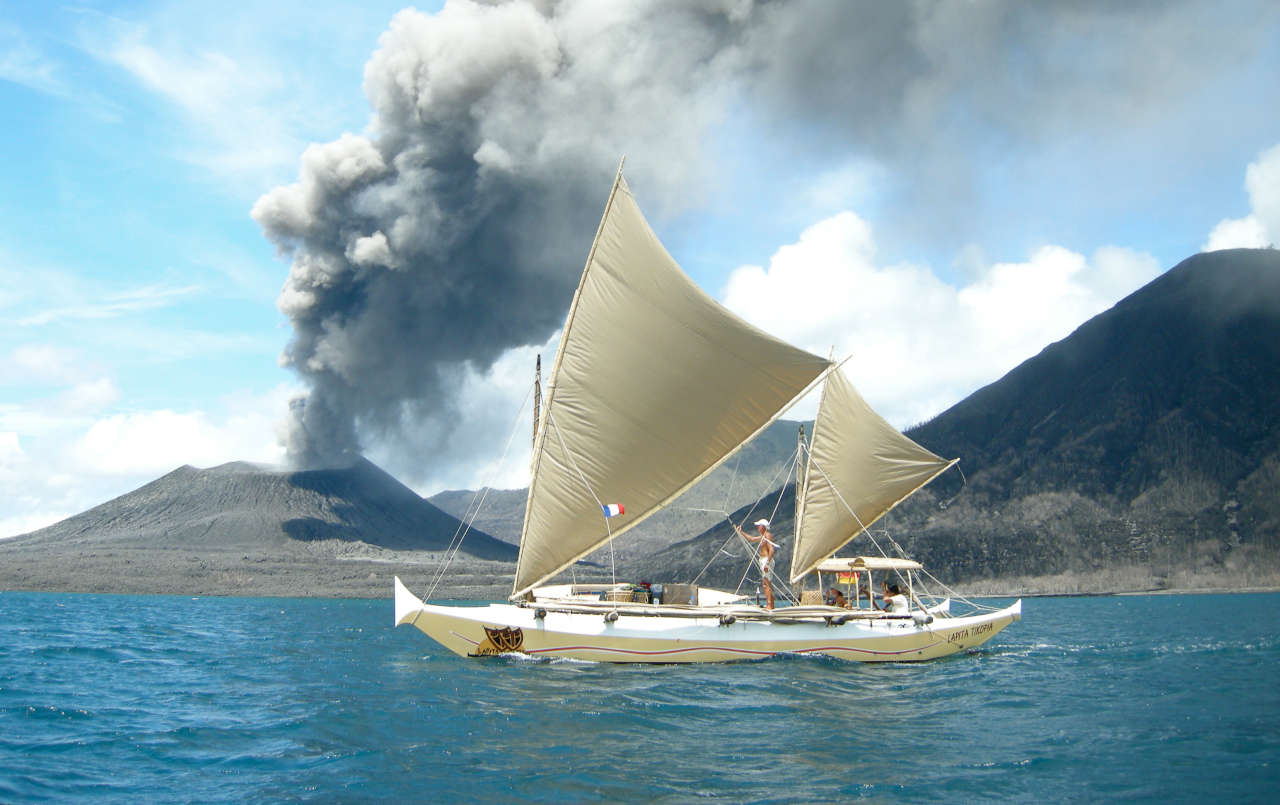 Double canoe catamaran sailing past a smoking volcano