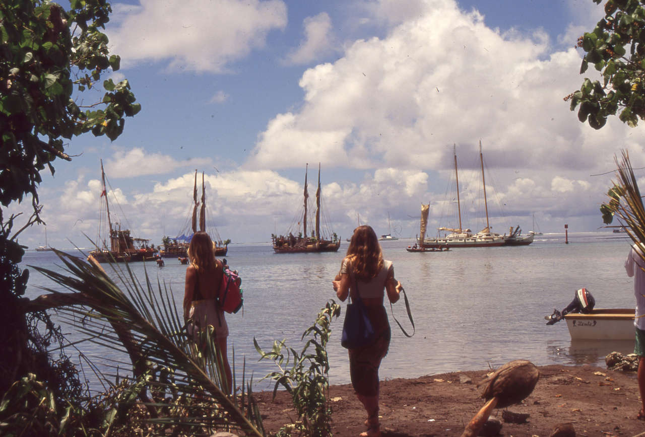 Two women looking out into a bay with various double canoe craft at anchor