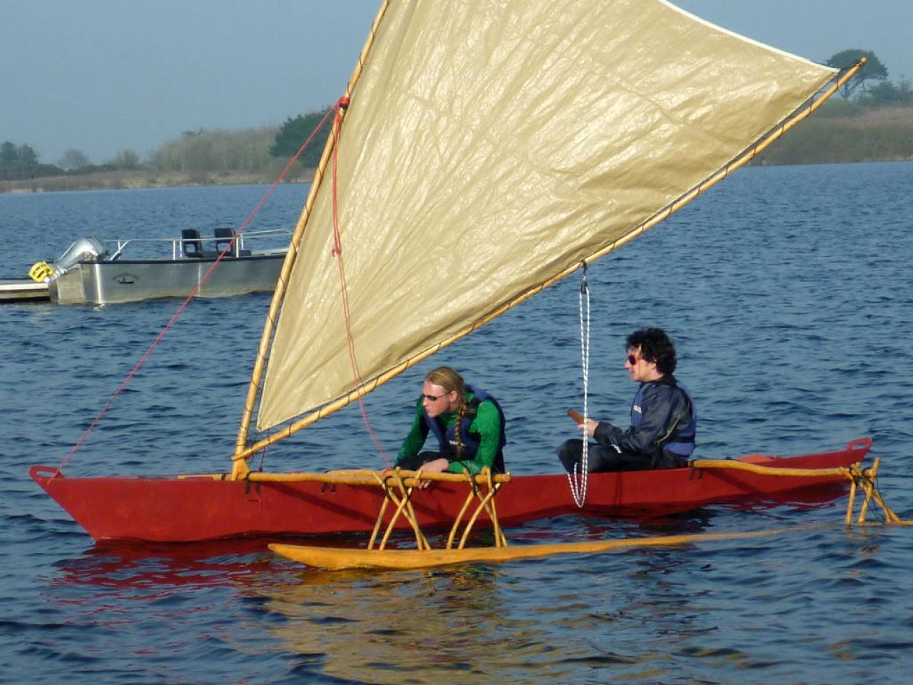 Small red outrigger canoe with crabclaw sail, two crew aboard